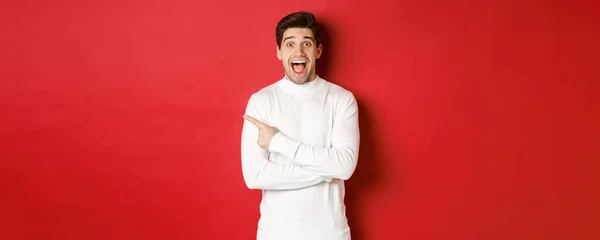 Concept of winter holidays, christmas and lifestyle. Amazed handsome man in white sweater showing logo, pointing finger left and looking excited, standing against red background — Stock Photo, Image