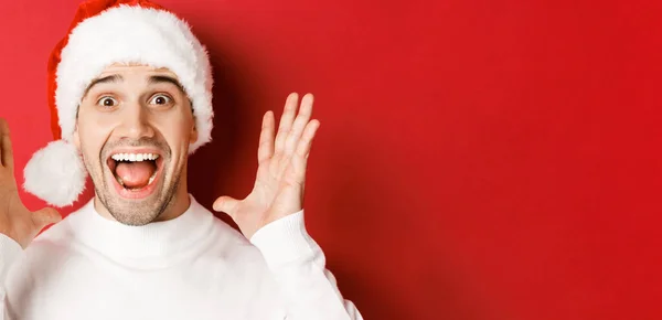 Close-up de jovem feliz em Papai Noel, fazendo grande anúncio de Natal, sorrindo espantado, de pé sobre fundo vermelho — Fotografia de Stock