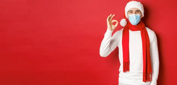 Concept covid-19, karácsony és ünnepek világjárvány idején. Portré boldog és elégedett férfi orvosi maszk és Mikulás kalap, mutatja rendben jel jóváhagyása, ajánló valamit — Stock Fotó