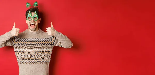 Imagem de cara bonito animado em óculos de festa e camisola de Natal, mostrando polegares para cima, elogiando grande celebração do ano novo, de pé sobre fundo vermelho — Fotografia de Stock