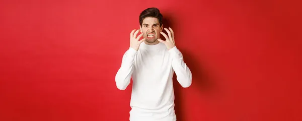 Image of angry and pissed-off man in white sweater, grimacing and shaking from rage, standing furious against red background — Stock fotografie