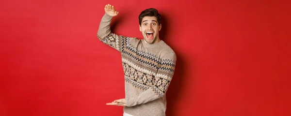 Portrét vzrušeného a šťastného pohledného muže ve vánočním svetru, ukazuje něco velkého, drží velký dárek na dovolenou, stojí nad červeným pozadím — Stock fotografie