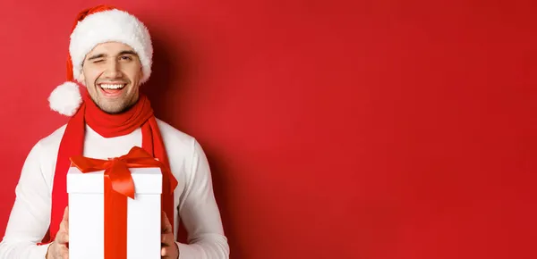 Kış tatili, Noel ve yaşam tarzı kavramı. Noel Baba şapkası ve eşarplı yakışıklı adam, hediyesini tutuyor ve gülümsüyor, kameraya göz kırpıyor, kırmızı arka planda duruyor. — Stok fotoğraf