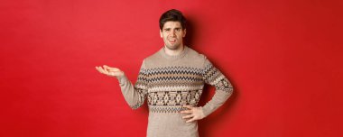 Kafası karışmış ve hayal kırıklığına uğramış yakışıklı bir adamın resmi, Noel partisinden şikayet eden, elini kaldıran ve rahatsız görünen, kırmızı arka planda duran