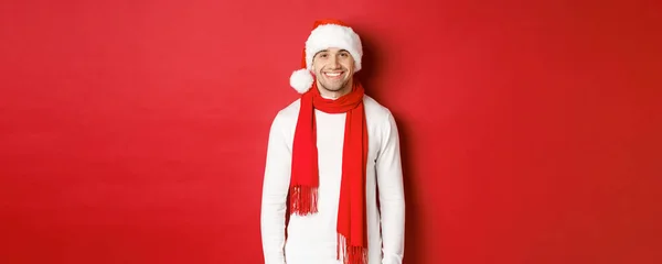 Cara caucasiano feliz comemorando o Natal e sorrindo, vestindo chapéu de Papai Noel, cachecol e suéter branco, desfrutando de feriados de ano novo, de pé sobre fundo vermelho — Fotografia de Stock