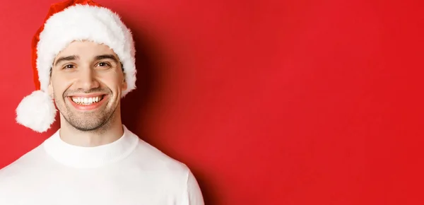 Primer plano del atractivo hombre sonriente en jersey blanco y sombrero de santa, luciendo feliz, disfrutando de las vacaciones de invierno, de pie sobre fondo rojo — Foto de Stock
