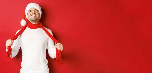 Beeld van verontruste en gefrustreerde man in santa hoed, wurgend zichzelf met sjaal van verdriet, staande tegen rode achtergrond — Stockfoto