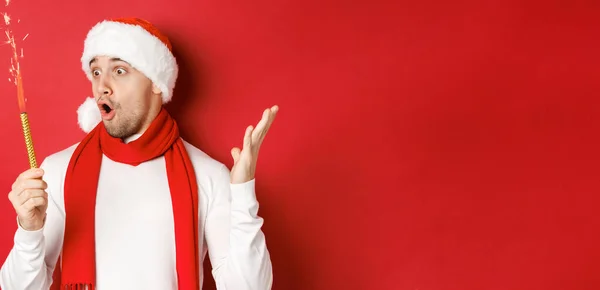 Koncepcja Bożego Narodzenia, ferie zimowe i uroczystości. zbliżenie zdumiewający przystojny mężczyzna w Santa kapelusz i szalik, patrząc na blask z podnieceniem, stojąc na czerwonym tle — Zdjęcie stockowe