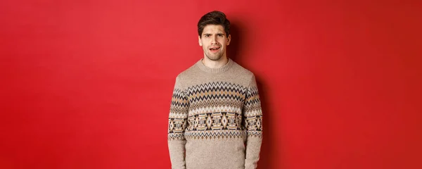 Porträt eines verwirrten erwachsenen Mannes im Weihnachtspulli, der enttäuscht im Neujahrsurlaub steht, etwas nicht versteht, vor rotem Hintergrund steht — Stockfoto
