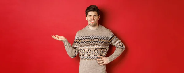 Image of confused and disappointed handsome man, complaining about christmas party, raising hand and looking bothered, standing over red background — Stock Photo, Image