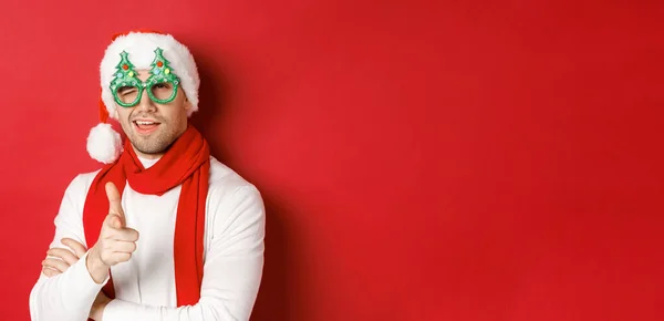 Concept de Noël, vacances d'hiver et célébration. Gros plan d'un jeune homme effronté dans un chapeau de Père Noël et des lunettes de fête, souriant et pointant du doigt pistolet à la caméra, debout sur fond rouge — Photo