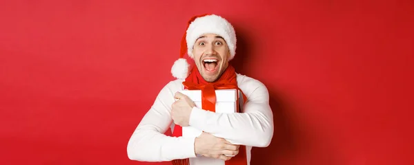 Концепция зимних праздников, Рождества и образа жизни. Крупный план счастливого человека в шляпе Санты, получающего подарок, выглядящего счастливым и обнимающим подарочную коробку, стоящего на красном фоне — стоковое фото