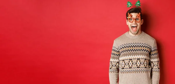 Изображение удивленного и взволнованного привлекательного мужчины, носящего очки для вечеринок с рождественским свитером, опущенной челюстью и смотрящего на удивительное новогоднее промо-предложение, стоящего на красном фоне — стоковое фото