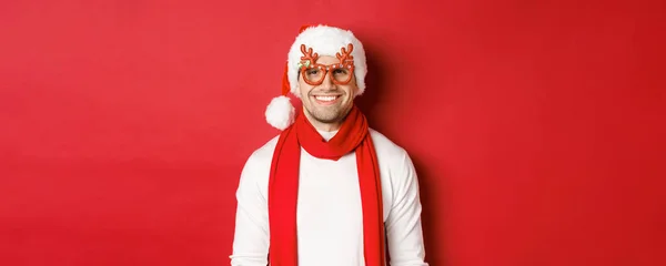 Concept de Noël, vacances d'hiver et célébration. Joyeux beau gars dans les lunettes de fête de nouvel an et souriant, debout sur fond rouge — Photo