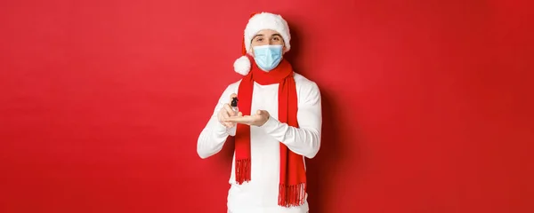 Covid-19, noel ve salgın sırasında tatil kavramı. Noel Baba şapkalı ve tıbbi maskeli mutlu genç adam, ellerini antiseptikle temizliyor ve gülümsüyor, kırmızı arka planda duruyor. — Stok fotoğraf