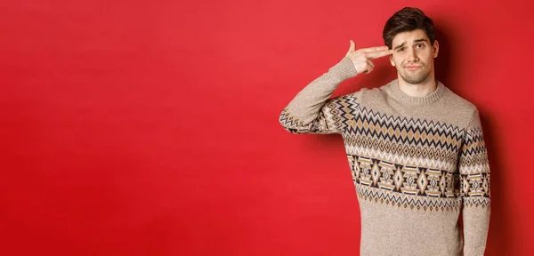 Retrato de homem bonito angustiado e irritado, vestindo camisola de Natal, fazendo dedo arma sinal perto da cabeça e atirando em si mesmo, de pé sobre fundo vermelho — Fotografia de Stock