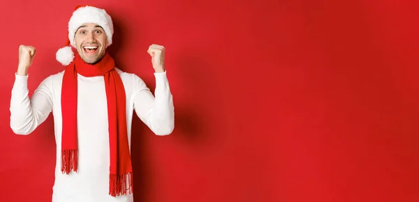 Porträt eines glücklichen und aufgeregten Mannes mit Weihnachtsmütze und Schal, der sich freut und etwas gewinnt, Neujahr feiert, vor rotem Hintergrund steht — Stockfoto