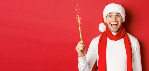 Begrip van Kerstmis, wintervakantie en viering. Knappe man die nieuwjaar viert en plezier heeft, een ster vasthoudt en glimlacht, een kerstmuts draagt, over een rode achtergrond staat — Stockfoto