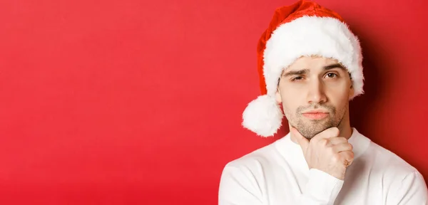 Close-up van bedachtzame knappe man met kerstmuts, fronsen en naar de camera kijken, aan iets denken, over een rode achtergrond staan — Stockfoto
