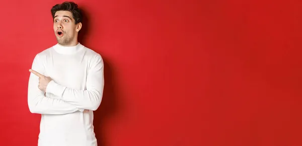 Concept of winter holidays. Portrait of impressed handsome man in white sweater, pointing and looking left with dropped jaw and amazed expression, standing against red background — Stock Photo, Image