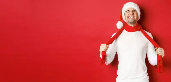 Noel Baba şapkalı sıkıntılı ve hayal kırıklığına uğramış bir adamın resmi. Üzüntü eşarbıyla kendini boğuyor, kırmızı arka planda duruyor. — Stok fotoğraf