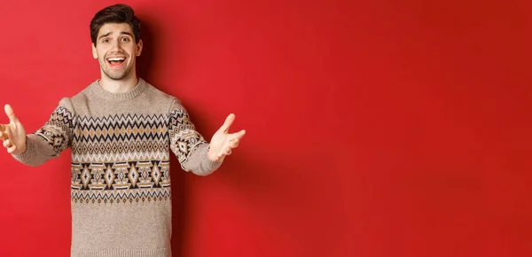 Image of happy and flattered handsome guy in christmas sweater, reaching hands to welcome guests on new year party, inviting to celebration, standing over red background — Stock Photo, Image