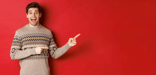 Concept of christmas celebration, winter holidays and lifestyle. Handsome man in xmas sweater pointing fingers right, smiling amazed, showing new year promo against red background — Stock Photo, Image