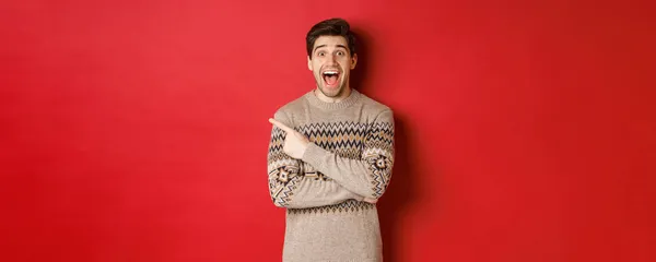 Portré csodálkozó jóképű srác, visel karácsonyi pulóver, ujjal mutogatott a bal felső sarokban logó és izgatott, mutatja félelmetes újévi promo ajánlat, felett áll piros háttér — Stock Fotó