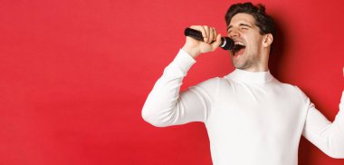 Beyaz kazaklı yakışıklı bir adam, şarkı söylüyor, mikrofonu tutuyor ve karaoke barda sahne alıyor, kırmızı arka planda duruyor.