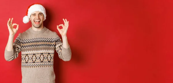Imagem de cara atraente alegre em suéter e chapéu de santa, desejando Feliz Natal, mostrando sinais ok e piscando para a câmera, celebrando o ano novo, de pé sobre o fundo vermelho — Fotografia de Stock