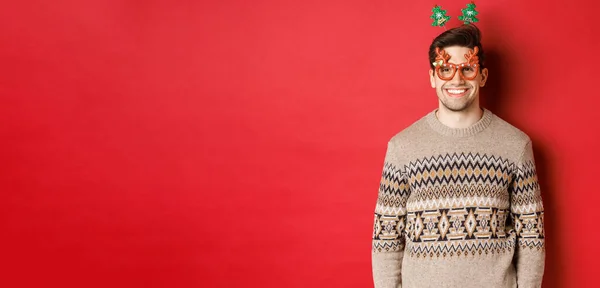 Porträtt av stilig skäggig kille i festglasögon och vinter tröja, ler glad, firar nytt år, önskar god jul, står över röd bakgrund — Stockfoto