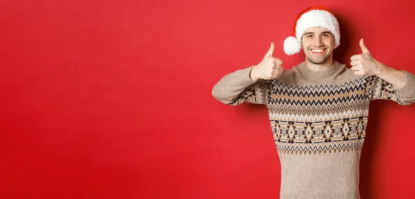 Portret atrakcyjnego szczęśliwego mężczyzny w kapeluszu i swetrze Mikołaja, z kciukami do góry w aprobacie i uśmiechem, życząc wesołych Świąt, stojąc nad czerwonym tłem — Zdjęcie stockowe