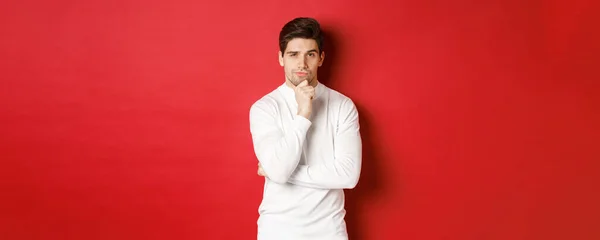 Image of thoughtful handsome man making assumption, thinking and looking at camera, standing in white sweater against red background — Stock Photo, Image