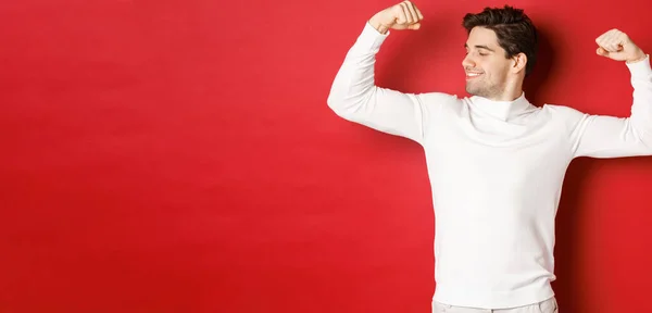 Retrato de hombre guapo sonriente en suéter blanco, bíceps flexionantes y jactancia con fuerza, músculos fuertes después del entrenamiento, de pie sobre fondo rojo —  Fotos de Stock