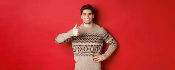Retrato de hombre guapo feliz en suéter de Navidad, sonriendo y mirando satisfecho, mostrando pulgares hacia arriba en la aprobación, como vacaciones de año nuevo, de pie sobre el fondo rojo — Foto de Stock