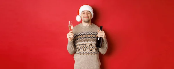 Konzept der Winterferien, Weihnachten und Feiern. Bild eines zufriedenen lächelnden Mannes mit Weihnachtsmütze und Pullover, der an Neujahr trinkt, eine Flasche Champagner und ein gefülltes Glas in der Hand hält, roter Hintergrund — Stockfoto