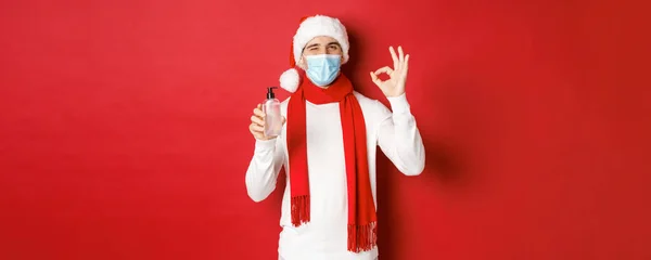 Концепція ковіда-19, Різдво та свята під час пандемії. Веселий чоловік, який святкує новий рік в медичній масці і капелюсі Санта, рекомендує дезінфікуючий засіб для рук, показуючи добре знак і підморгнувши — стокове фото