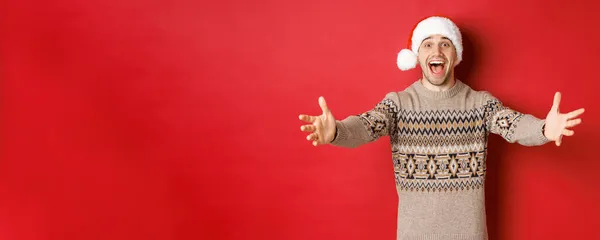 クリスマスのセーターとサンタの帽子の幸せ、魅力的な男の肖像は、贈り物のための手に達し、何かを取りたいと笑顔、赤い背景の上に立って — ストック写真