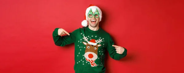 Образ щасливого чоловіка в партійних окулярах і капелюсі Санти, вказуючи на його різдвяний светр і посміхаючись, стоїть на червоному тлі — стокове фото