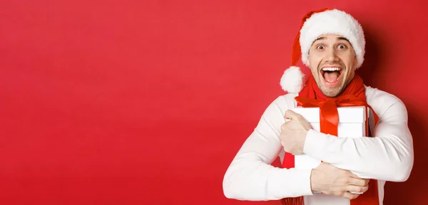 Concept de vacances d'hiver, Noël et style de vie. Gros plan de l'homme heureux dans le chapeau de Père Noël recevant cadeau, regardant heureux et étreignant boîte cadeau, debout sur fond rouge — Photo