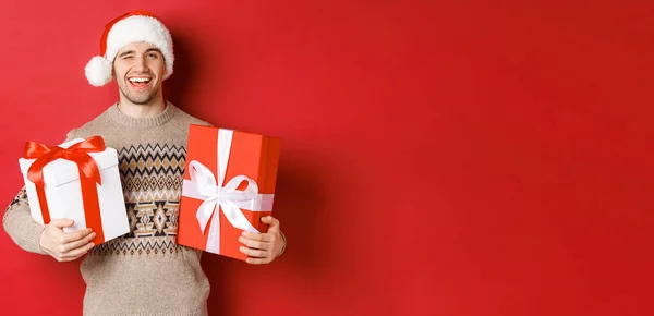 Koncepcja ferii zimowych, Nowy Rok i świętowanie. Portret pewnego siebie i bezczelnego młodego człowieka przygotował prezenty na Boże Narodzenie, mrugając i trzymając prezenty, stojąc na czerwonym tle — Zdjęcie stockowe