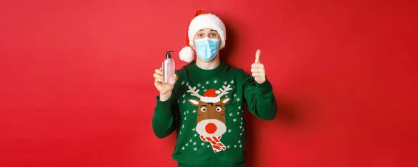 Έννοια του νέου έτους, coronavirus και κοινωνική αποστασιοποίηση. Πορτρέτο του χαρούμενου άνδρα σε καπέλο Σάντα, χριστουγεννιάτικο πουλόβερ και ιατρική μάσκα, που δείχνει τον αντίχειρα-up και συνιστώντας απολυμαντικό χεριών — Φωτογραφία Αρχείου