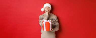 Noel Baba şapkalı ve Noel süveterli yakışıklı bir adam resmi, Noel hediyesini açmak için heyecanlı, şaşırmış görünüyor ve hediyeyi bir elinde tutuyor, kırmızı arka planda duruyor.