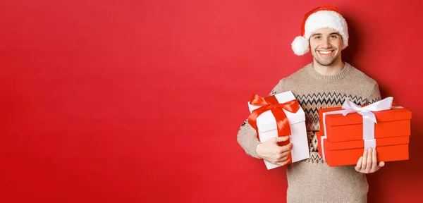 Konzept der Winterferien, Neujahr und Feier. Porträt eines gutaussehenden Mannes mit Weihnachtsmütze und Pullover, der Schachteln mit Weihnachtsgeschenken in der Hand hält und lächelt, vorbereitete Geschenke, roter Hintergrund — Stockfoto