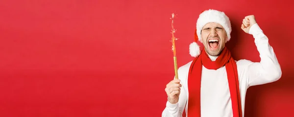 Konzept von Weihnachten, Winterurlaub und Feiern. Schöner Mann feiert Neujahr und hat Spaß, hält Wunderkerze in der Hand und lächelt, trägt Weihnachtsmütze, steht über rotem Hintergrund — Stockfoto