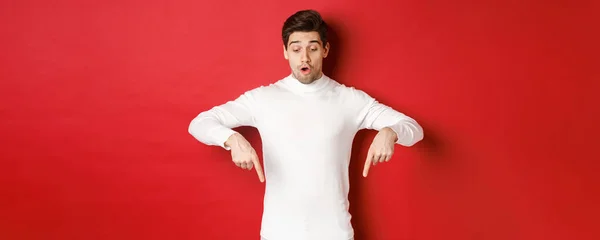 Portrait of surprised good-looking guy in white sweater, looking and pointing fingers down at logo, standing against red background — Stock Photo, Image