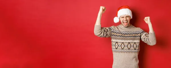 Zufriedener gutaussehender Mann, der sich glücklich und glücklich fühlt, vor Freude schreit und Faust-Pumps macht, Sieg oder Niederlage feiert, ein tolles Weihnachtsgeschenk erhält, im Weihnachtsmann-Hut vor rotem Hintergrund steht — Stockfoto