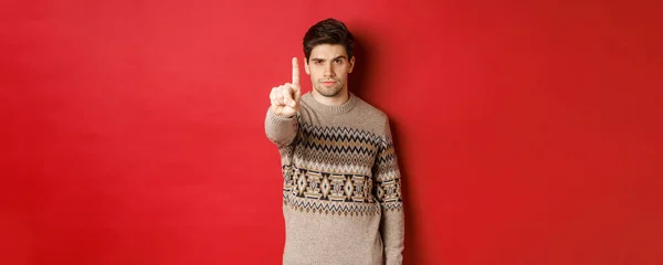 深刻なハンサムな男のイメージがあなたを止めるために1本の指を伸ばし、警告を与え、冬のセーターの赤い背景の上に立って — ストック写真