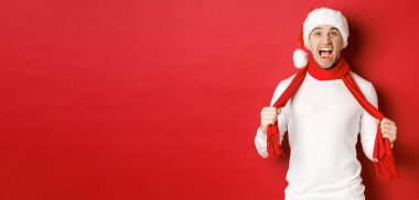 Kızgın yetişkin bir adamın resmi Noel 'den nefret eder, eşarp takar ve Noel Baba şapkası takar, sıkıntılı bağırır, kırmızı arka planda kızgın durur.