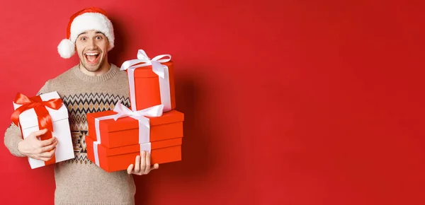 Conceito de férias de inverno, ano novo e celebração. Imagem de cara feliz desfrutando de Natal, segurando pilha de presentes e sorrindo divertido, de pé sobre fundo vermelho — Fotografia de Stock